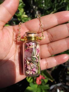 a hand holding a glass jar filled with pink and white flowers on a gold chain