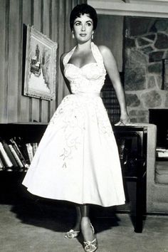 a woman in a white dress standing next to a book shelf with books on it