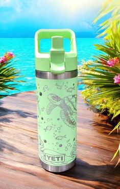 a water bottle sitting on top of a wooden table next to the ocean and flowers