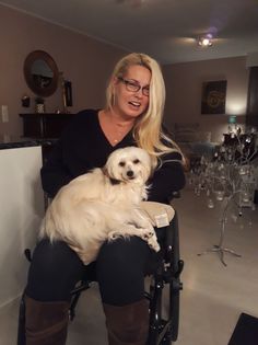 a woman sitting in a wheel chair holding a white dog with long blonde hair and brown boots