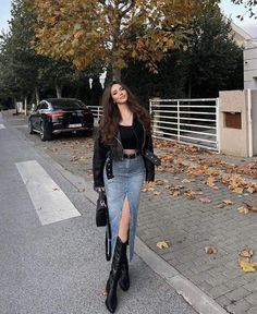 a woman standing on the side of a road next to a leaf covered tree and wearing a black top