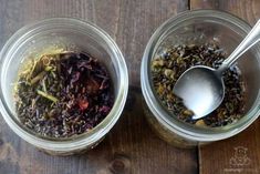 two glass jars filled with different types of tea on top of a wooden table next to a spoon