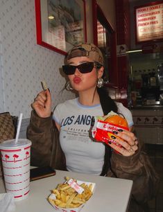 a woman sitting at a table with a sandwich and fries in her hand while holding a drink