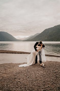 a bride and groom kissing on the shore of a lake with mountains in the background