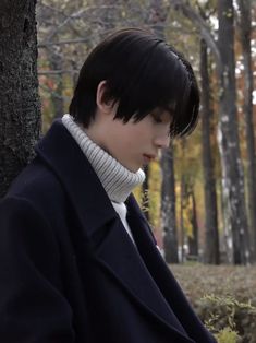 a young man sitting next to a tree looking at his cell phone while wearing a coat