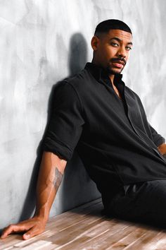 a man sitting on top of a wooden table next to a white wall with his hands in his pockets