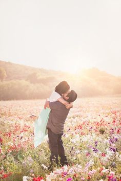 a man holding a woman in a field of flowers with the sun setting behind him