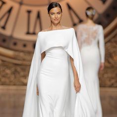 a model walks down the runway in a white gown with cape on her shoulders and long sleeves