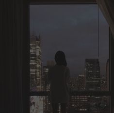 a woman standing in front of a window looking out at the city lights and skyscrapers