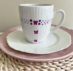 a white cup and saucer with purple flowers on it sitting on a pink plate