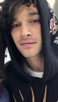 a man with long hair wearing a hoodie and looking at the camera while standing next to a woman