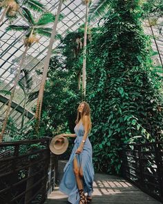 a woman in a blue dress and hat standing on a wooden walkway surrounded by palm trees