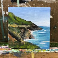 an acrylic painting of a cliff by the ocean on a wooden table with paintbrushes