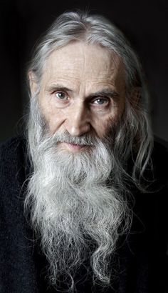 an old man with long white hair and beard wearing a black sweater is looking at the camera