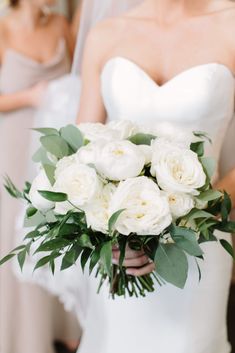 a bride holding a bouquet of white roses and greenery in front of two other bridesmaids