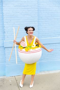 a woman in a yellow dress holding two wooden sticks and a large white bowl with flowers on it