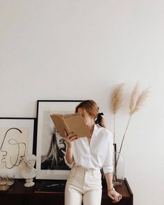 a woman standing in front of a white wall holding an open book and looking at it