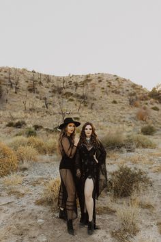 two women dressed in black standing next to each other on a dirt field with dry grass