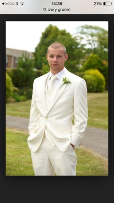 a man wearing a white suit and tie standing in front of a green lawn with trees