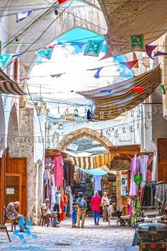 people are walking through an open market with colorful flags hanging from the buildings above them