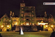 a bride and groom standing in front of an old castle at night with lights on