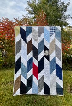 a quilt is hanging on the grass in front of some trees and bushes with red, white, blue, grey and black colors