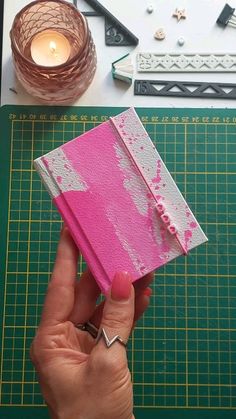 a person is holding up a pink piece of paper on a cutting board with other crafting supplies