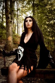 a woman in a black dress and white mask sitting on a bench with trees behind her