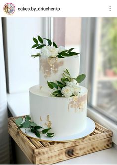 a three tiered cake with white flowers and greenery on the top is sitting in front of a window