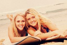 two beautiful young women laying on top of a surfboard in the sand at the beach