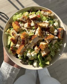 a person holding a bowl of salad with chicken and feta cheese on it's side