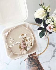 a person holding up a cake in a white box with flowers on the inside and writing'9 months '