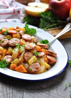 a white plate topped with sausage and vegetables next to apples on a wooden table,