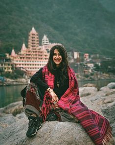 a woman sitting on top of a large rock next to a body of water with buildings in the background