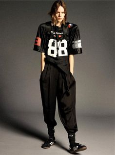 a woman standing in front of a gray background wearing black pants and a shirt with the number 86 on it