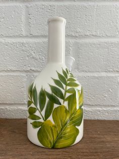 a white vase with green leaves painted on the front and sides, sitting on a wooden table