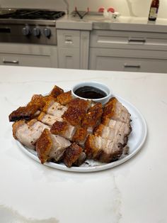 a white plate topped with sliced meat next to a cup of sauce on top of a counter