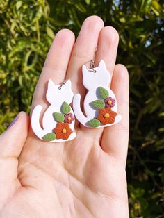 a hand holding two white cat shaped earrings with flowers on the front and back of them