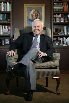 an older man in a suit and tie sitting in a chair with bookshelves behind him