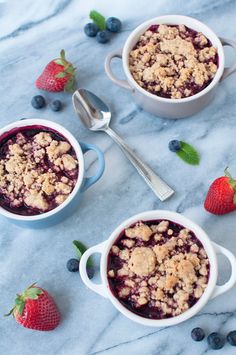 two white bowls filled with blueberries and crumbled oatmeal next to strawberries