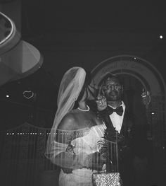 a bride and groom standing in front of a gate at night with lights behind them