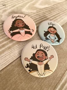 three buttons with cartoon characters on them sitting on a bed, one has her arms out and the other is smiling