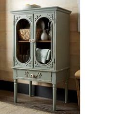 a green cabinet with two glass doors and baskets on it's top shelf, in front of a wood paneled wall