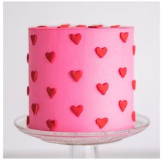 a pink cake with red hearts on it sitting on a glass stand, ready to be eaten