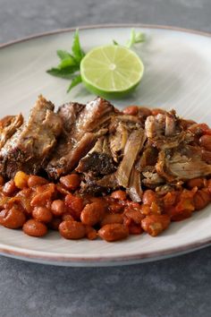 a white plate topped with meat covered in beans and garbanzo beans next to a lime wedge