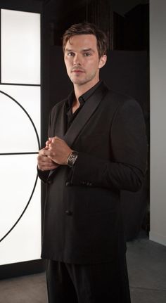 a man standing in front of a wall with a watch on his left hand and wearing a black suit