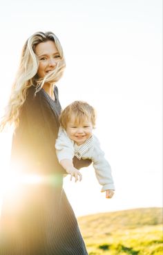 a woman holding a baby in her arms and smiling at the camera while standing on a field