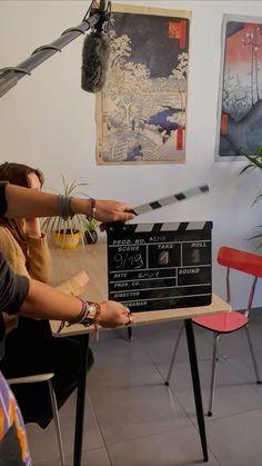 two people holding up a movie clapper in front of a table with chairs and pictures on the wall