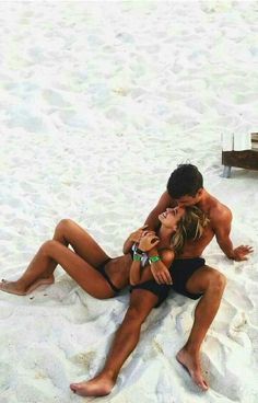 a man and woman are sitting on the beach in their bathing suits, one is kissing