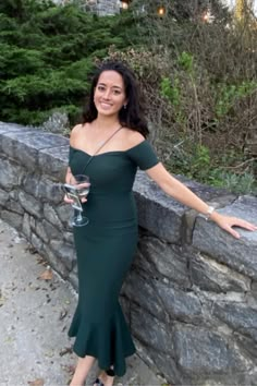 a woman in a green dress leaning against a stone wall holding a glass of wine